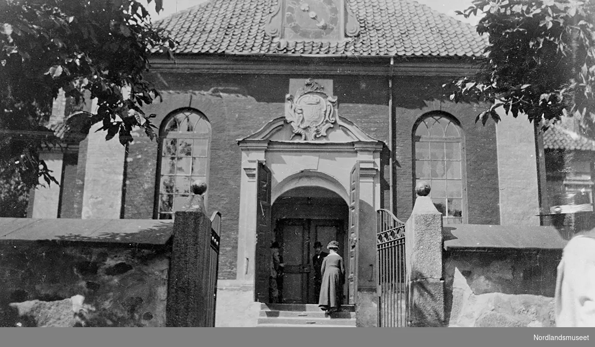 Stavern kirke, med ornament over døren, en klokke på taket. Midt på bildet tre personer ved inngangen, og en dame i mørk kåpe og hatt på vei opp trappen til inngangen. En steinmur med smijernsport i forgrunnen.