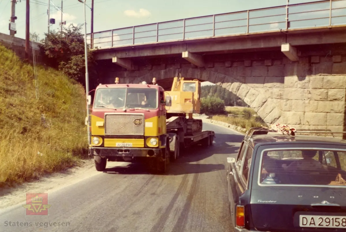 Rv.7 i 1976 gjennom Hønefoss kryss med jernbanen. Gammel og ny tid. 
Volvo lastebil 1970 modell  transporterer  Brøyt beltegraver. Registreringsnr. JU28490.
Bilen er en Ford 17 m Turnier (tredørs stasjonsvogn). Registreringsnr. DA29158.