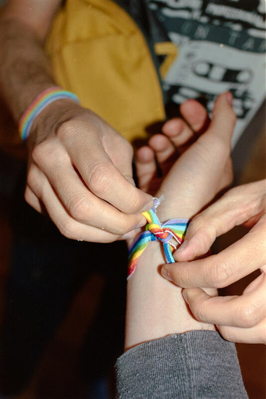 Bilde av hendene til en som knytter et pride armbånd på en armen til en annen person.