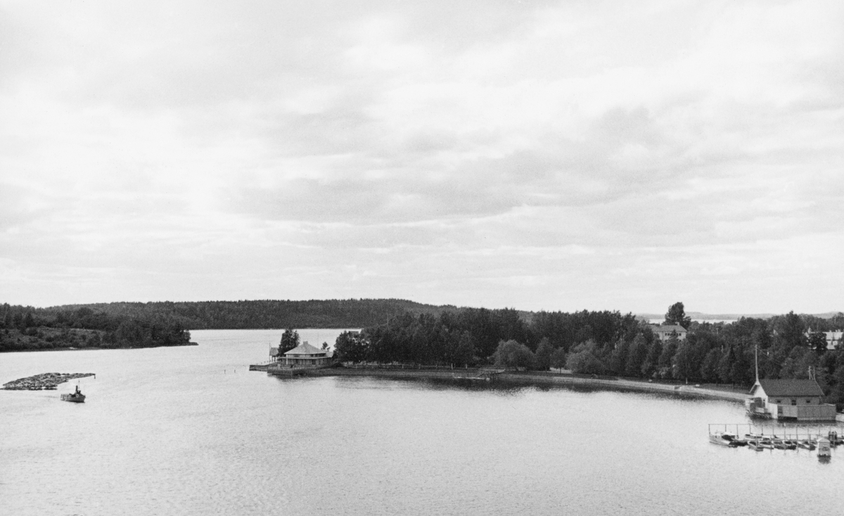 Innsjøfløting i Heinola i  Päijänne-Tavastland (Päijät-Häme) i Finland i 1934. Fotografiet er tatt fra et utsiktspunkt - visstnok ei jernbanebu - ned mot en innsjø. Til venstre i bildet ser vi et fartøy med et etterhengt tømmerslep med kurs innover i bildet. Til høyre, på motsatt side av innsjøen eller sundet, lå det ei havn for fritidsbåter og et badehus. På en odde sentralt i bildet ser vi en parkrestaurant.