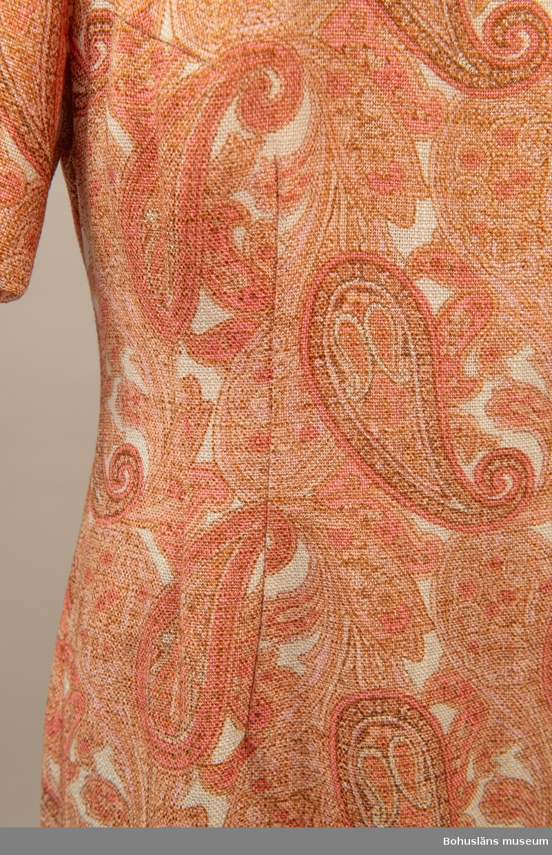 Rak fodrad klänning i så kallat persiskt mönster. Milda rosa färger.
Insvängd i midjan och dragkedja bak.  Se bilaga.

Ärmlängd 10cm.