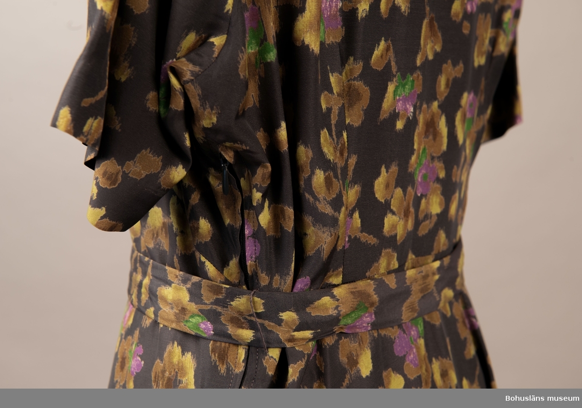 Klänning i konstsilkematerial med guld lila och grönt, blomster på lilagrå botten. Klockad kjol kort ärm och stor krage. Knäpps med tre knappar fram. Hör ihop med jacka UM.19787 i samma tyg som även syns på bild 1-3 samt 9-10.

Kjollängd 66cm.