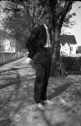 Portrett i helfigur av ung mann som står på en gårdsvei. Hus