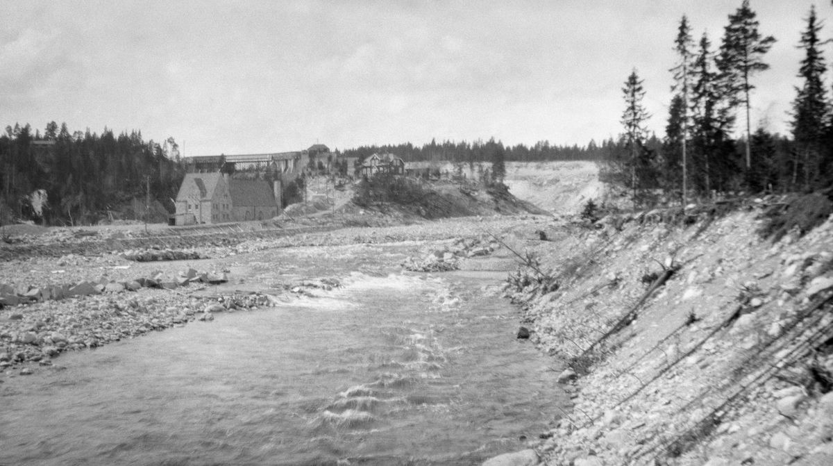 Fra den nedre delen av elva Søndre Osa i Åmot kommune i Østerdalen. Dette fotografiet er tatt i motstrøms retning, med en utrast elveskråning til høyre i forgrunnen. I bakgrunnen ser vi kraftstasjonen, bestyrerboligen og dammen, samt det nye elveløpet elva grov seg da den nordre fangarmen til dammen sviktet under en flom 10. mai 1916. Dette fotografiet ble tatt i 1923.