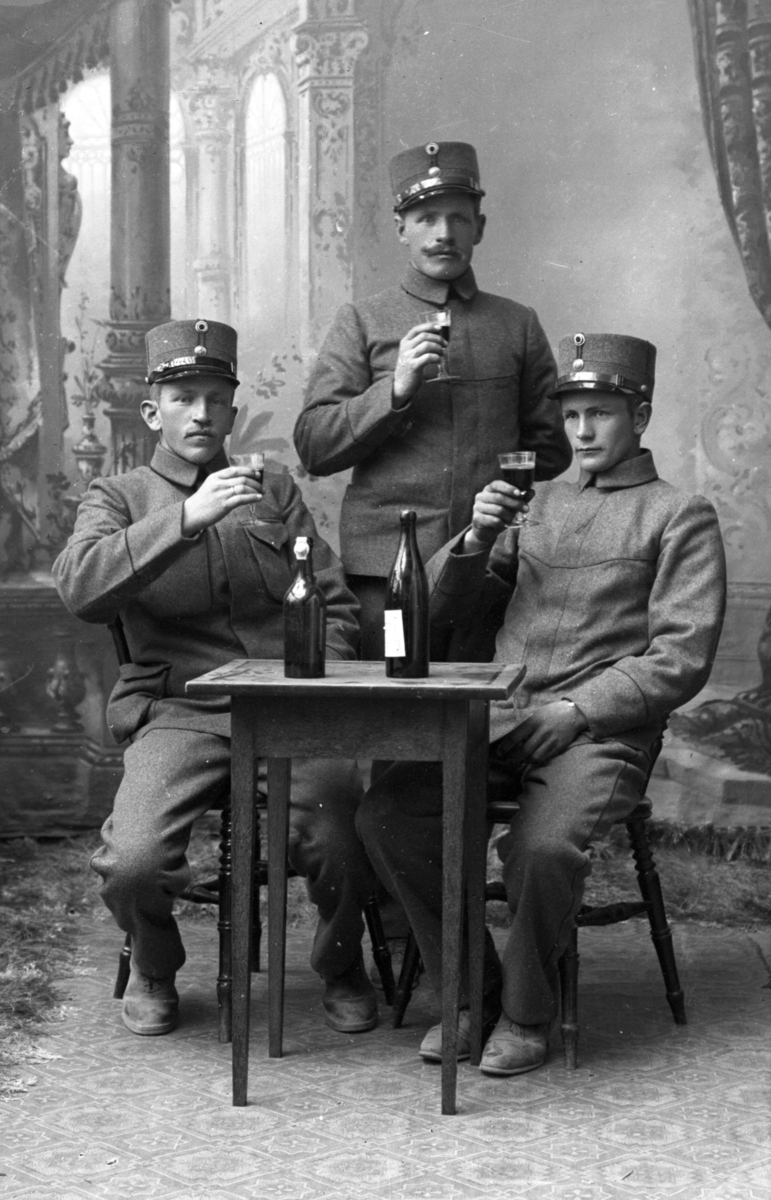Atelierfoto.Tre soldater sitter ved bord med vinflasker på og drikker vin.