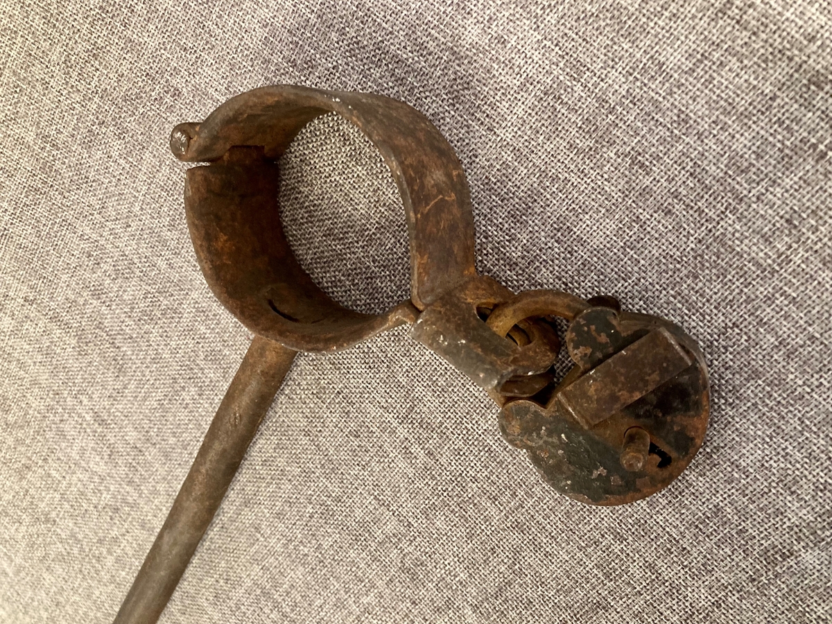 To leddede håndjern forbundet med en lang jernbolt. Begge jernene kan låses med hengelås. Den ene hengelåsen mangler.
