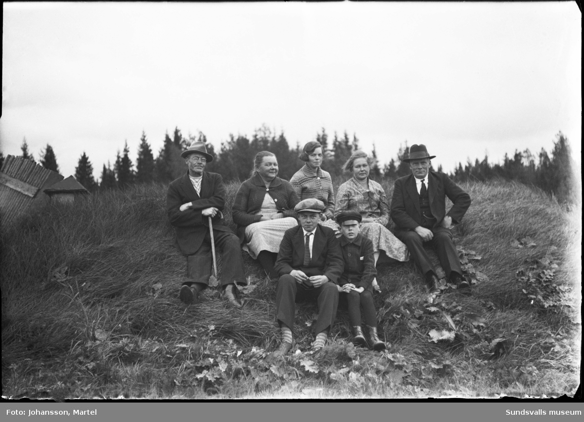 Gruppbild med barn och vuxna som sitter på en gräskulle. Troligen familjefoto.