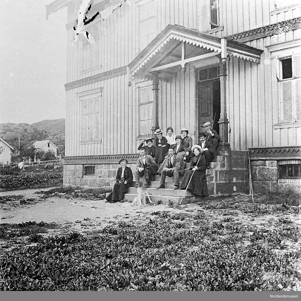 Bildet viser en gruppe mennesker som sitter på en trapp opp til inngangen til telegrafstasjonen i Lødingen. Foran sitter en hund. Det står en rive inntil veggen. Huset har overbygg over trappa, med utsmykninger. I bakgrunnen til venstre skimtes andre bygninger og noen trær.