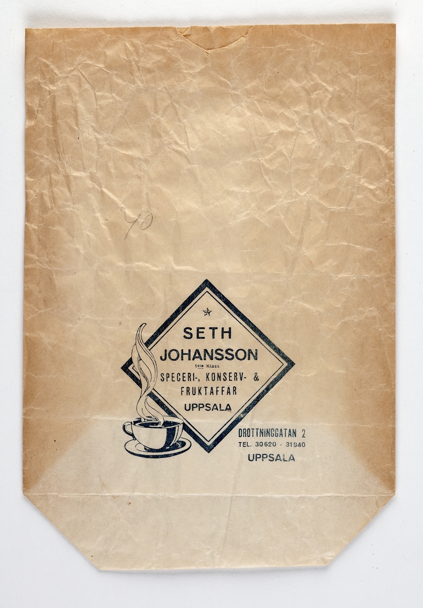 Beige delvis blekt påse. På framsidan blått tryck en ruta med text samt en kaffekopp. text: "Seth Johansson speceri-, konserv- & fruktaffär, Uppsala" samt texten "Drottninggatan 2, tel. 30620 - 31940, Uppsala"