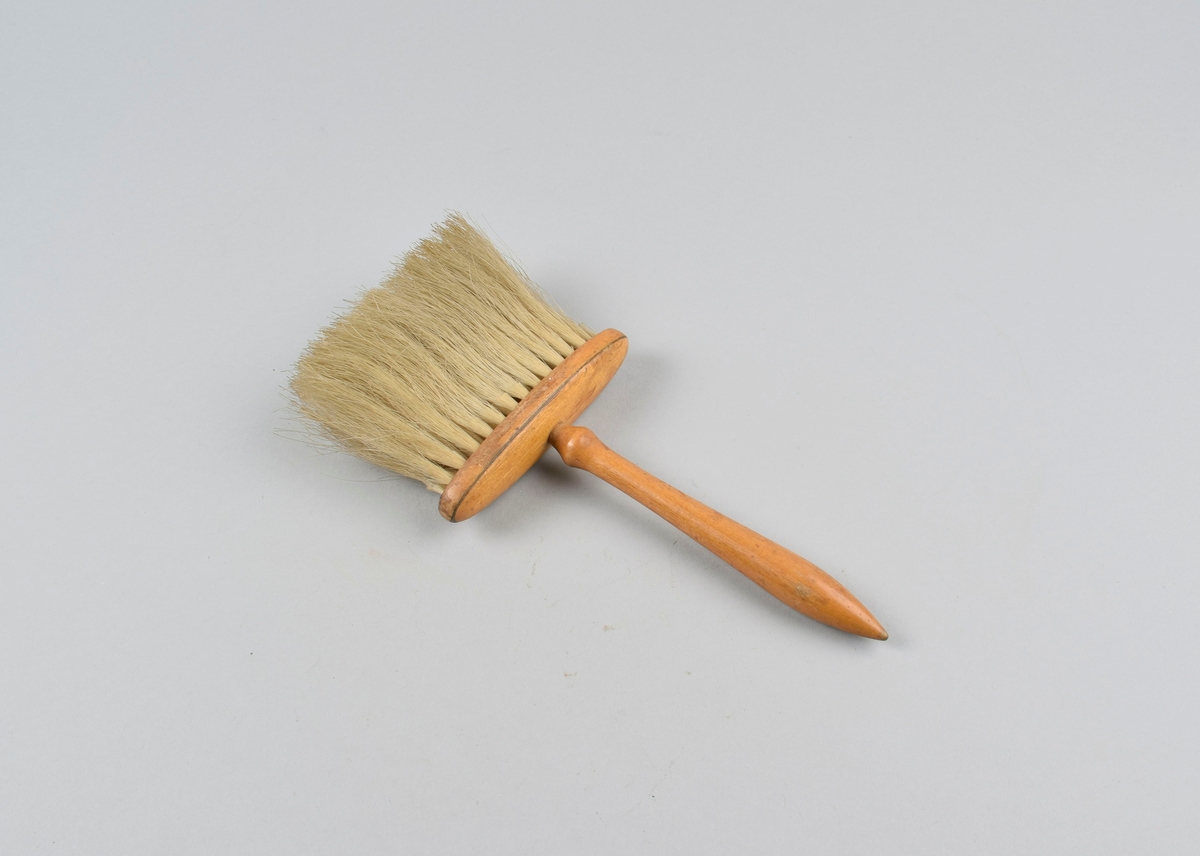 Flat børste med lange buster festet til oval plate med dreiet håndtak.
