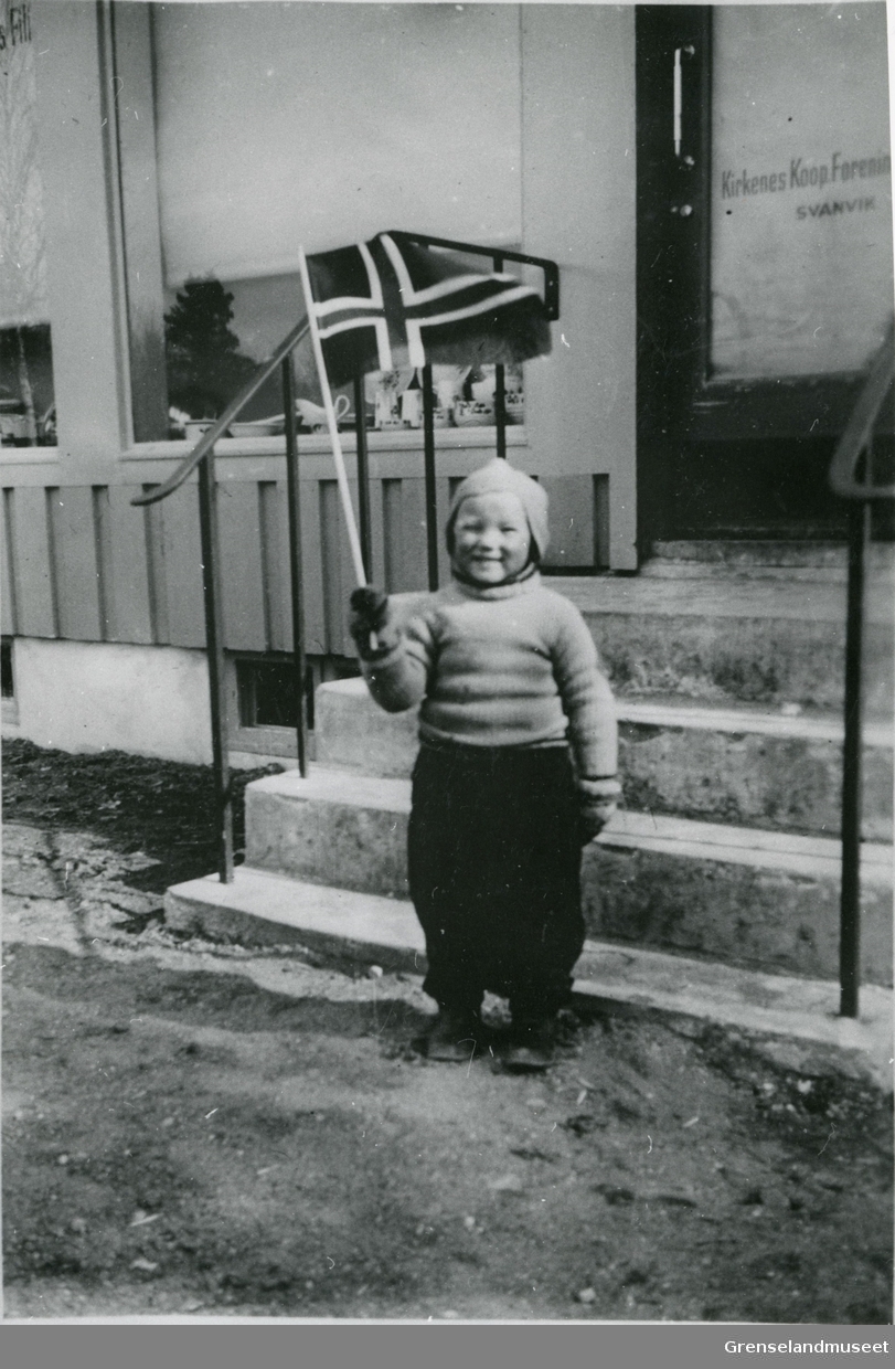 En av sønnene til Sterk (mulig Hans) står med et norsk flagg i hånden utenfor "Kirkenes Koop Forening" på Svanvik. 