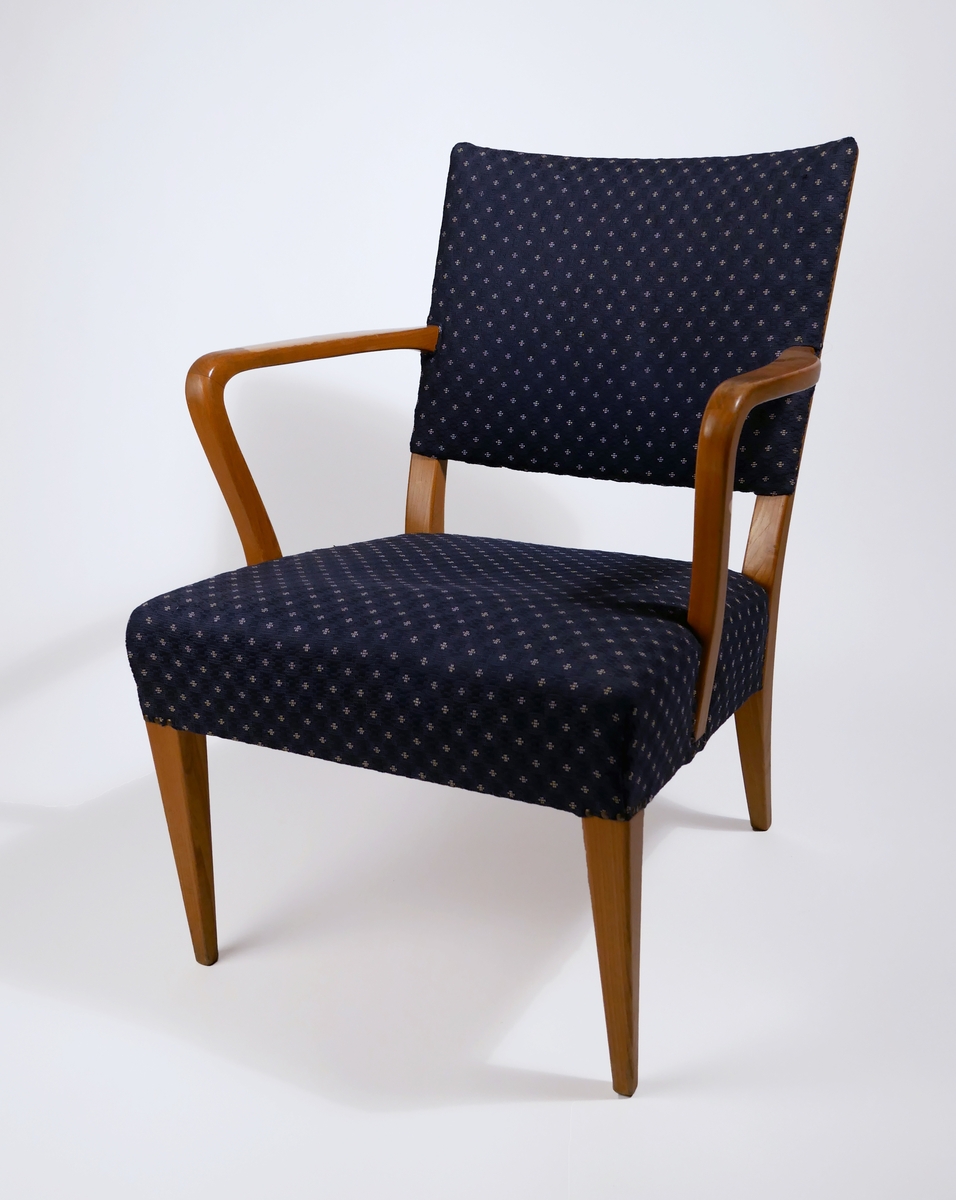 Fåtölj med stoppad sits och rygg, klädd med blått tyg. Fåtöljen kommer från Axelssons snickeri där den formgavs av Baltzar Axelsson 1942, tillverkades av Bertil Svensson och stoppades och kläddes av Svante Bergfelt.