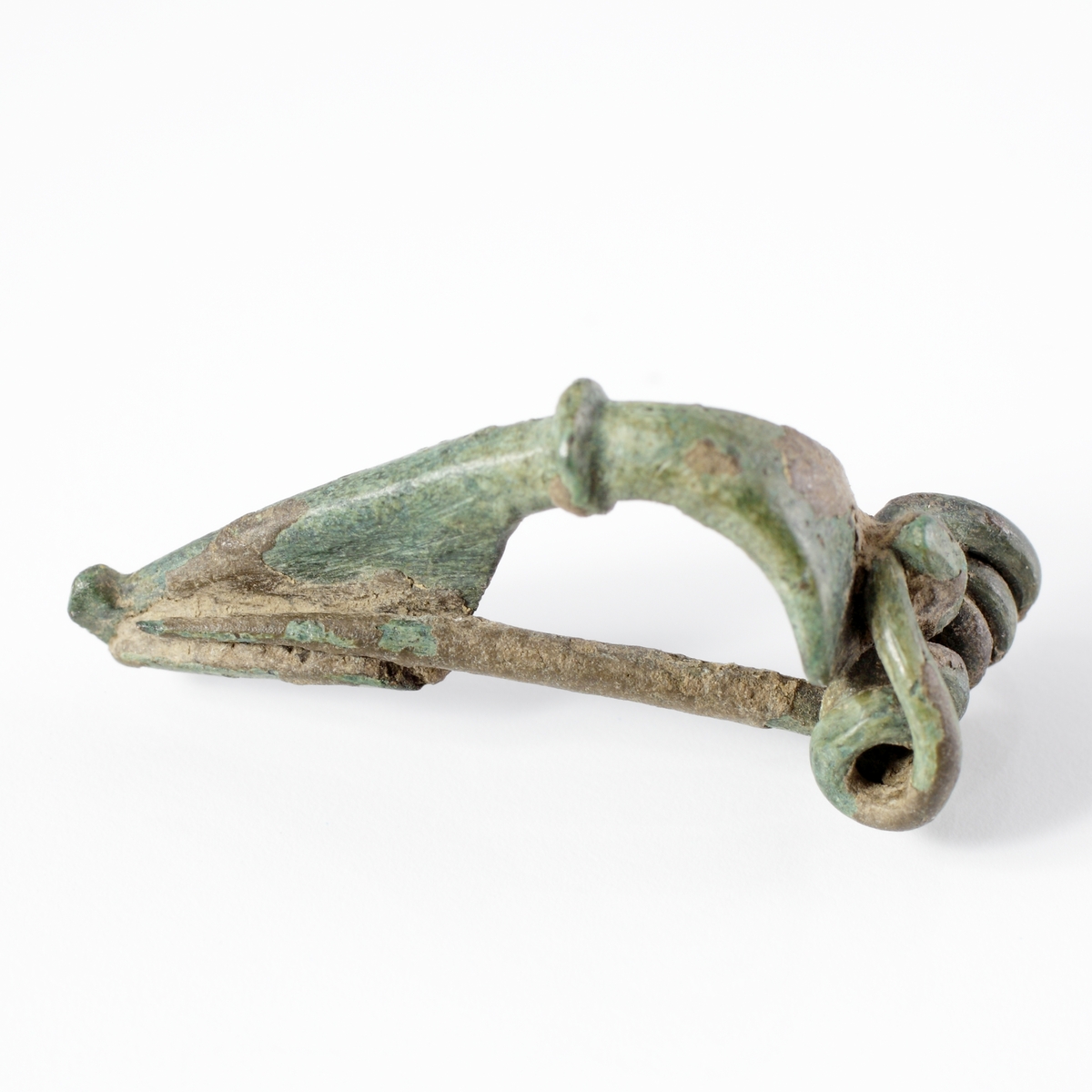 Trekantfibula av  brons med en liten vulst mellan båge och fot. Spiralfjädern fäster med en krok i änden på bågen.