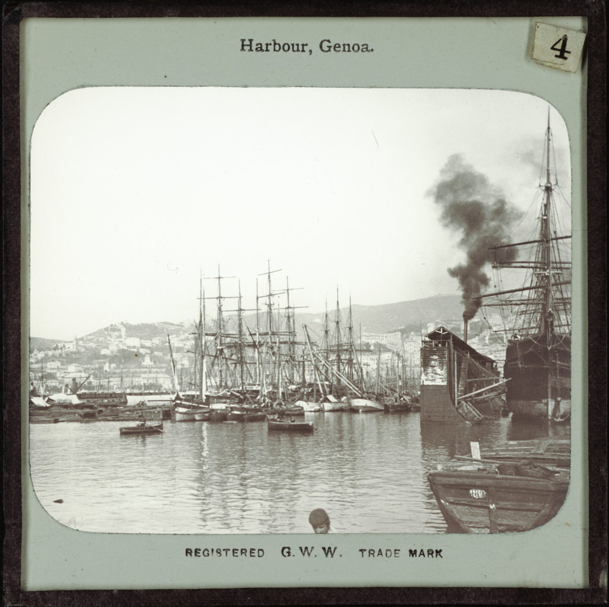 Bild från Italien föreställande hamnen Genua, "Harbour, Genoa". I hamnen finns enbart segelfartyg, men tjock svart rök, kanske från ett ångfartyg, stiger upp bakom en av de utskjutande pirerna. I bilden mitt syns två roddbåtar.
Registered G.W.W. Trade Mark.