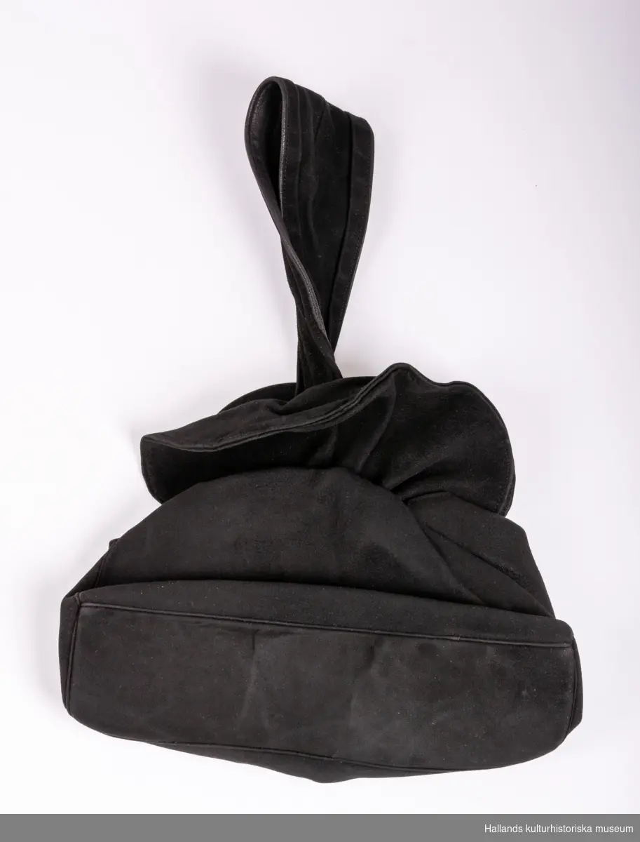Handväska i svart mocka, svart sidenfoder. Rektangulär svart botten, styv. Raka sidor som samlas likt en påse medelst ett mässingsspänne. Ovankanten rundad och rynkad. Handtag i form av fastsydd ögla.