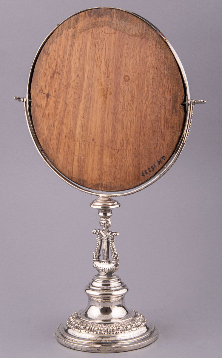 Bordsspegel i silver och glas. Tillverkad av silversmed Arvid Öhrn i Gävle, 1841. Rund på fot. Infattning m.m. av silver. Spegeln är vridbar runt i det halvcirkelformiga fästet, baksidan trä, teak? Rikligt med bladornament i kraftig relief.
I foten lyra med människohuvud med pagefrisyr. 
Stämplad "AÖ, G, L4"