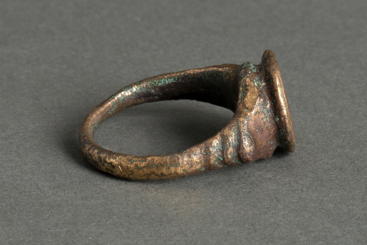 Støpt fingerring i messing(-legering) eller bronse. Ringen er smal og ujevn på baksiden, bredere mot forsiden og med profilert "skulderparti" på hver side av en sirkelrund plate med gravering: "AV".