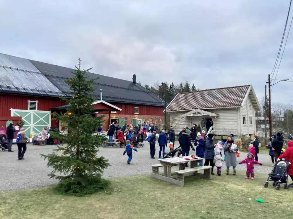 Et gårdstun med et hvitt stabbur og en rød låve, en stor folkemengde og et juletre