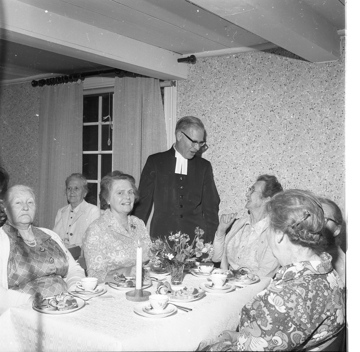 En präst står bland äldre kvinnor som sitter vid kaffebord, de samtalar och skrattar. Ett ljus är tänt på bordet.