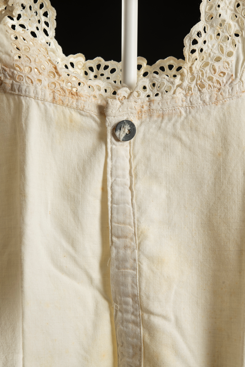 Kortärmad klänning av vitt vävt tyg. Spets runt ärm och halslinning. Bak ett 20 cm sprund med en knapp högst upp. Tyget är missfärgat på flera ställen.