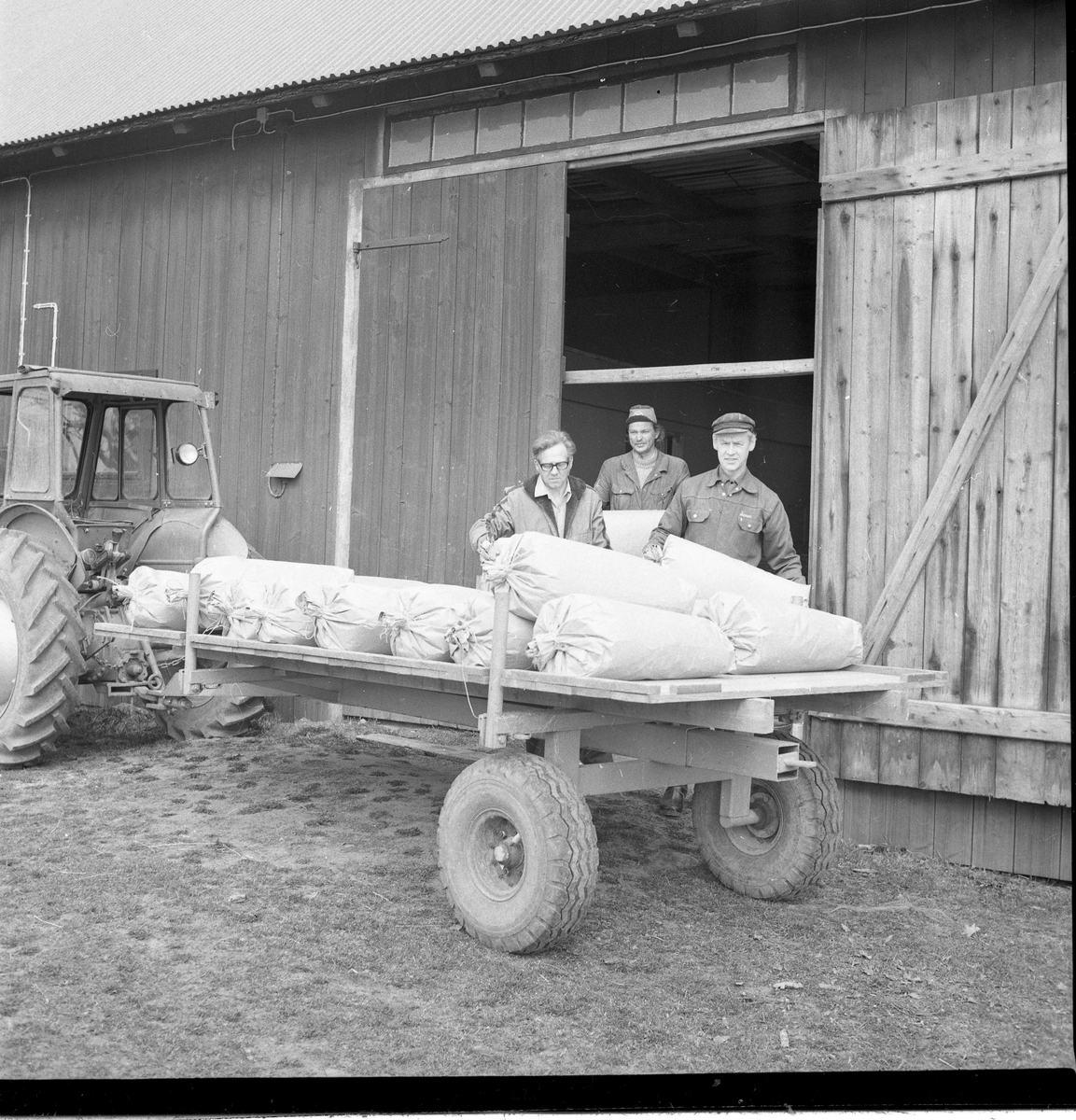 En flakvagn med säckar kopplad till en trakt står utanför en ekonomibyggnad, tre män lastar på mer säckar - troligen med müsli.