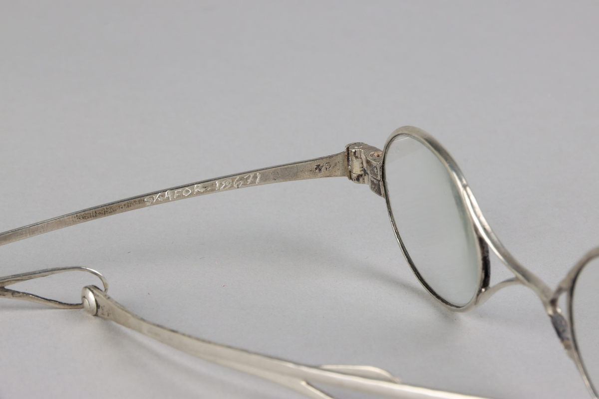 Silver oval eye spectacles, with hourglass shaped bridge and lenses. Sliding sides with small loops at the end for a riband. Hallmarks on front of slides.