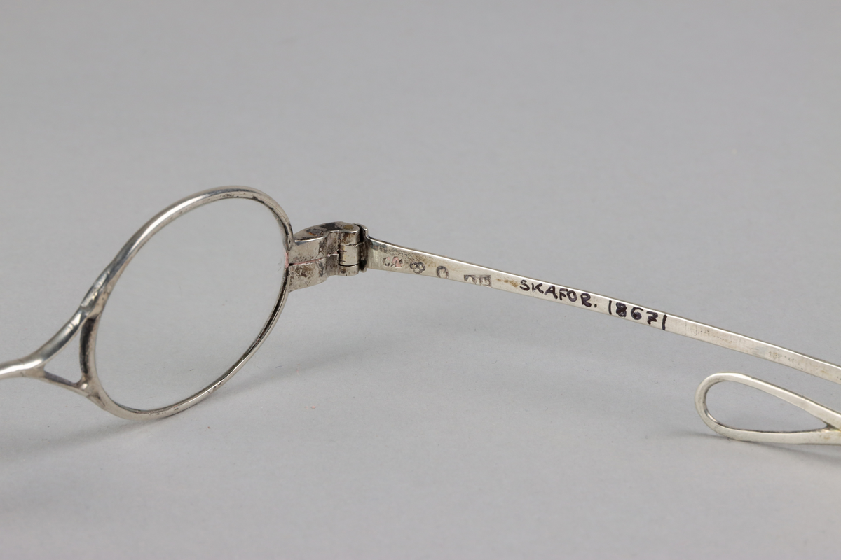 Silver oval eye spectacles, with hourglass shaped bridge and lenses. Sliding sides with small loops at the end for a riband. Hallmarks on front of slides.