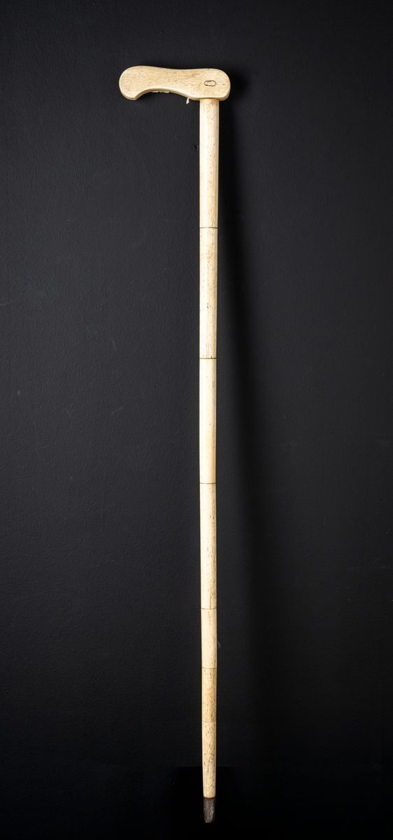 Spaserstokk av hvalbein med håndtak. Stokken er satt sammen av syv deler og med håndtak på toppen. Håndtaket er avlangt med en liten bøy. Tuppen er skodd med metall (de nederste 3cm).