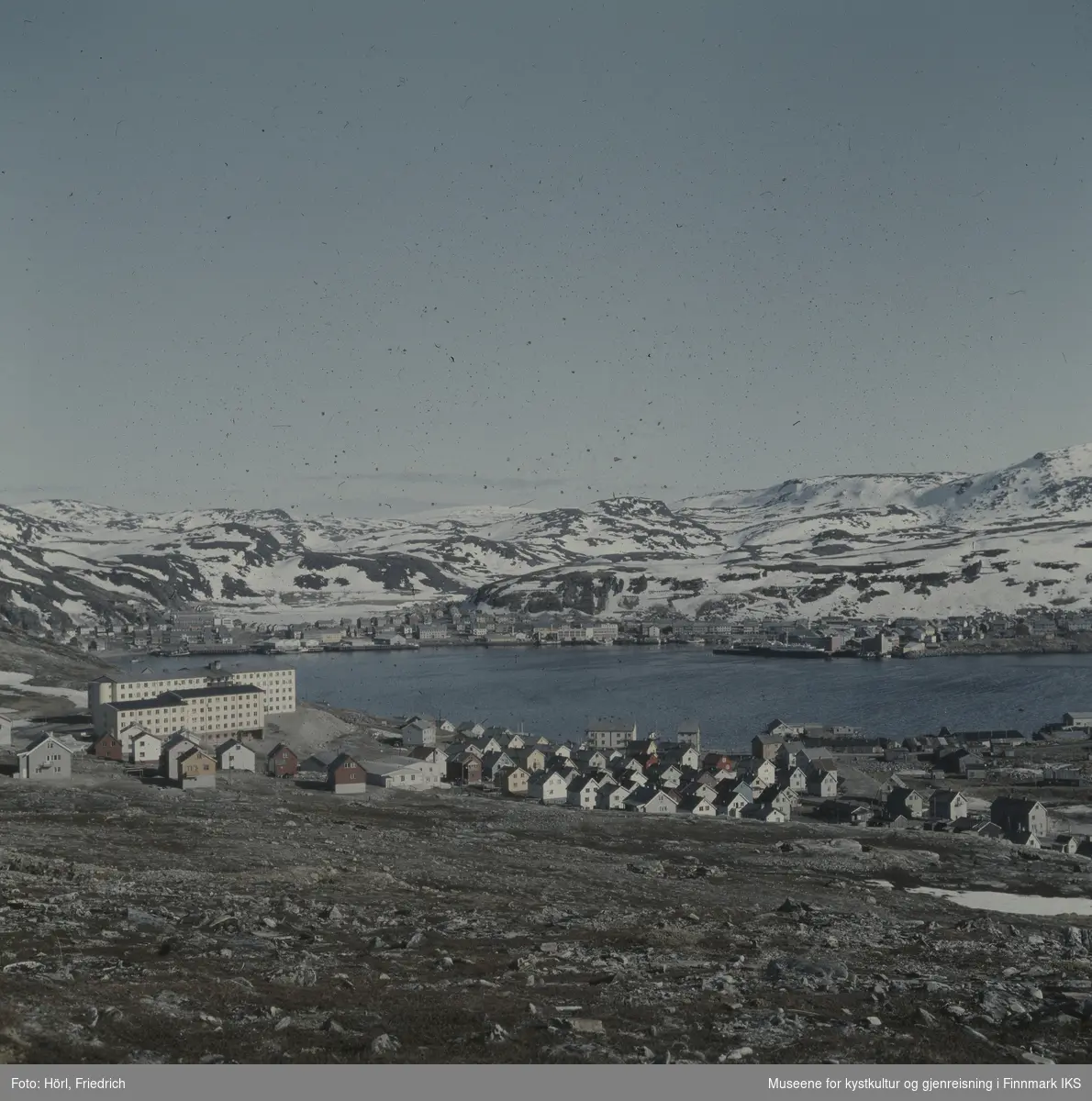 Utsikt over Hammerfest med sykehus og annet gjenreisningsbebyggelse i forgrunn. Bildet kan være tatt i våren 1958. Midt i bildet ser man havnebukta i Hammerfest, Mollaområde og byens sentrum bak den. På Storvannet og fjellene ligger det fortsatt snø.