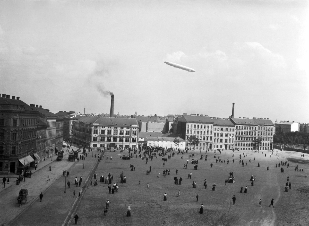Militärt luftskepp i luften över park i Wien i Österrike-Ungern. Människor i rörelse i parken. Vy från sidan.