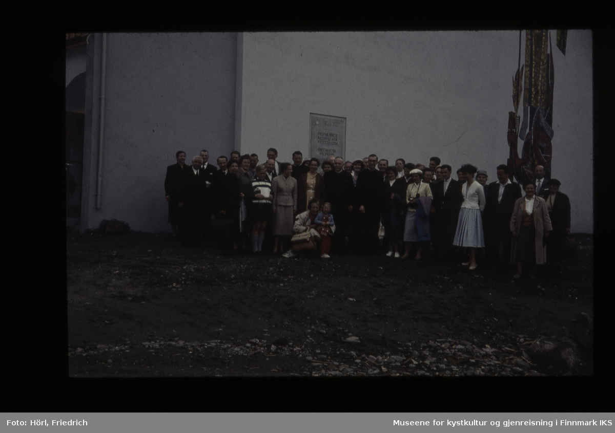 En gruppe personer i ulik alder står foran Den katolske kirke St. Mikael i Hammerfest i 1958. De er festkledde. Bak dem ser man deler av Karl Manningers veggmosaikk.