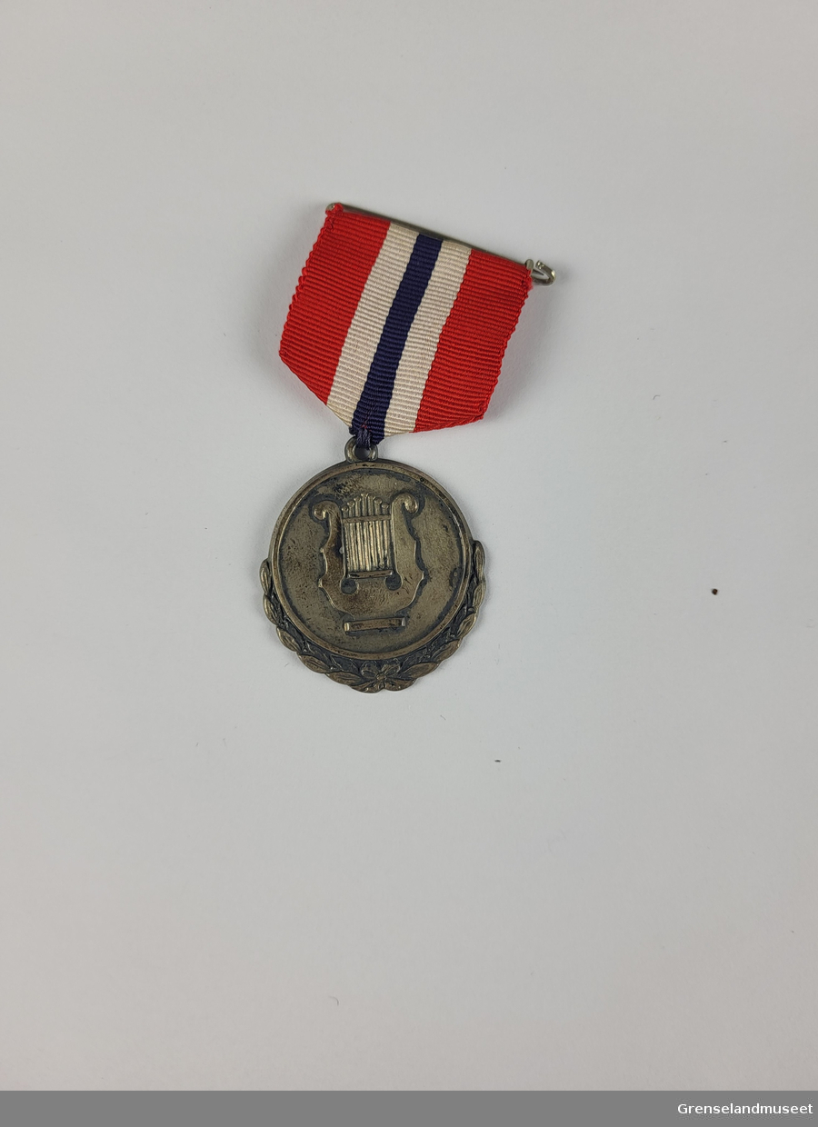 En lyre harpe i sølv uthevet fra resten av sølvmedaljen. Det norsk flaggets farger i tekstil som er festet til medaljen.  