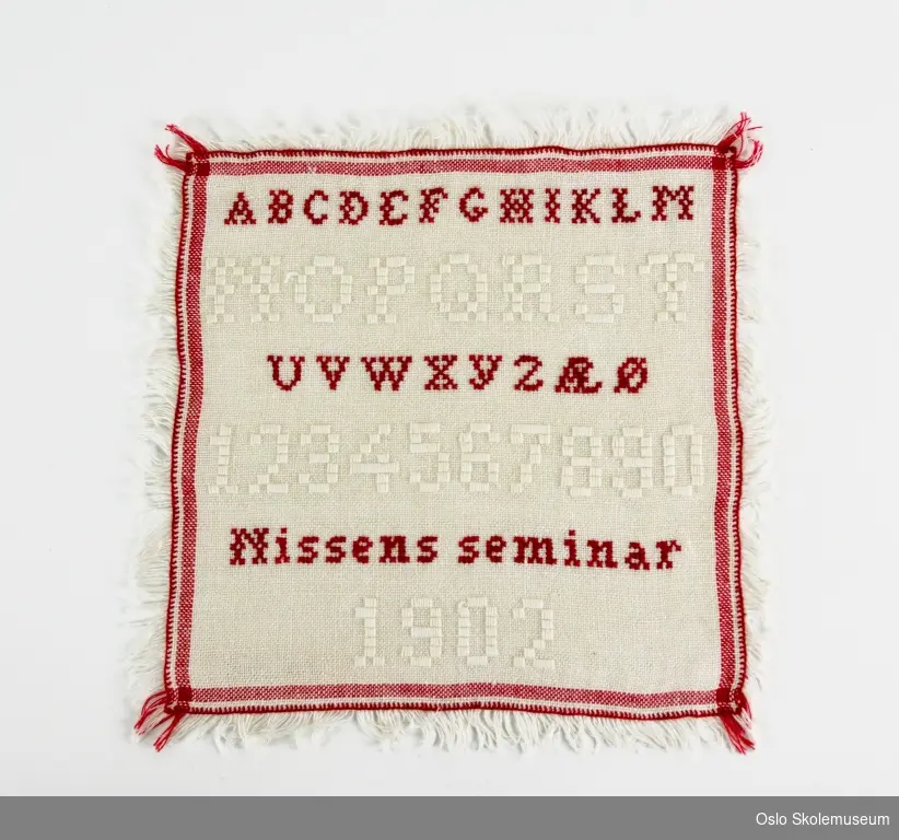 Kvadratisk navneduk med røde og hvite sting. På navneduken er det brodert bokstaver og tall samt "Nissens seminar" og "1902". Det er også brodert en pyntebord.