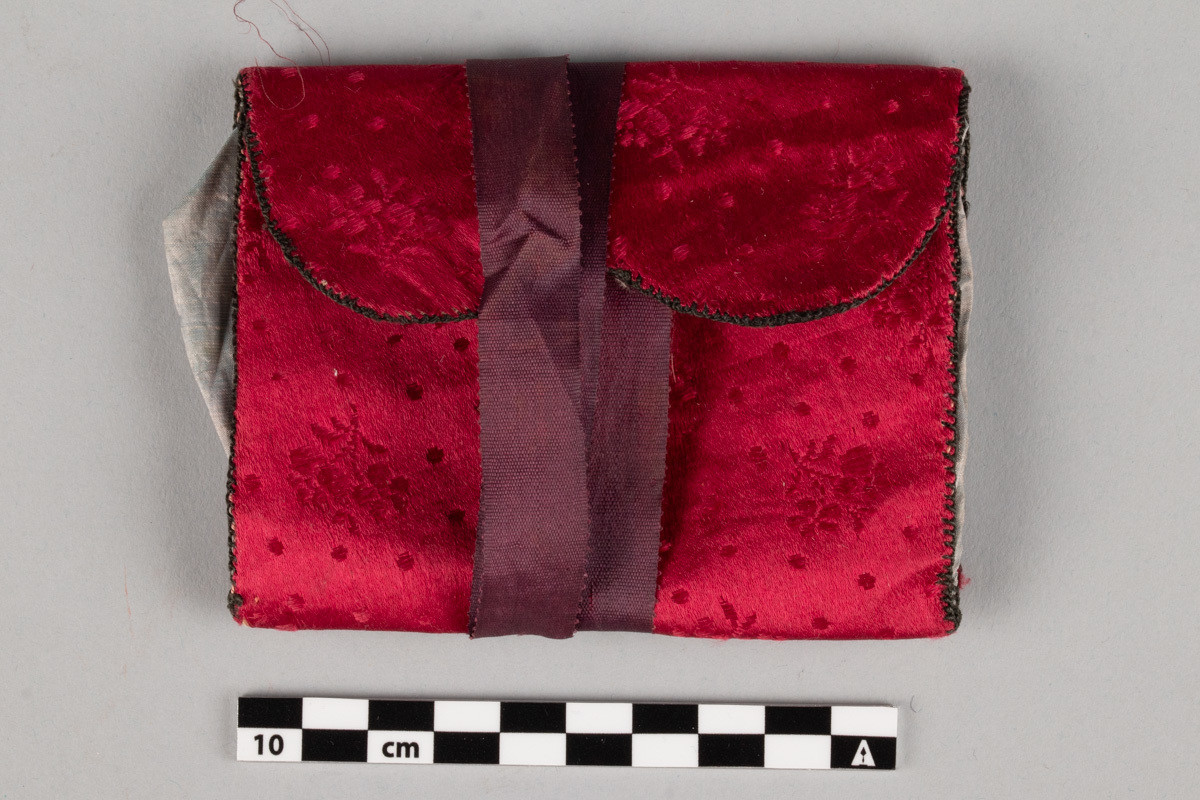 Rektangulær, etui av rød silke. Blomstermønster  vevd inn i silken. Svart innvendig. Grå lommevegger. Kan lukkes med lilla sløyfe bånd som surres rundt flere ganger