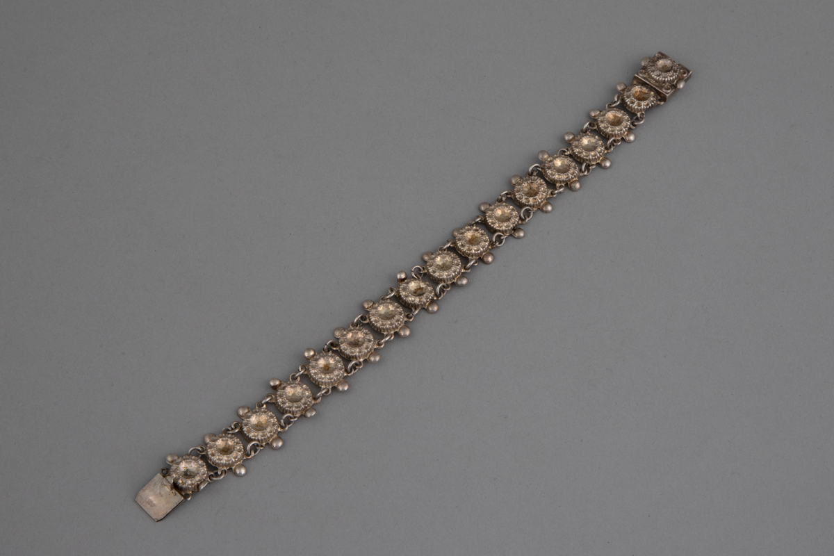 Halsbånd i sølv bestående av 16 like sammenlenkede ledd og kvadratisk lås. Leddene har form som rosetter med fasettslipt midtstykke.