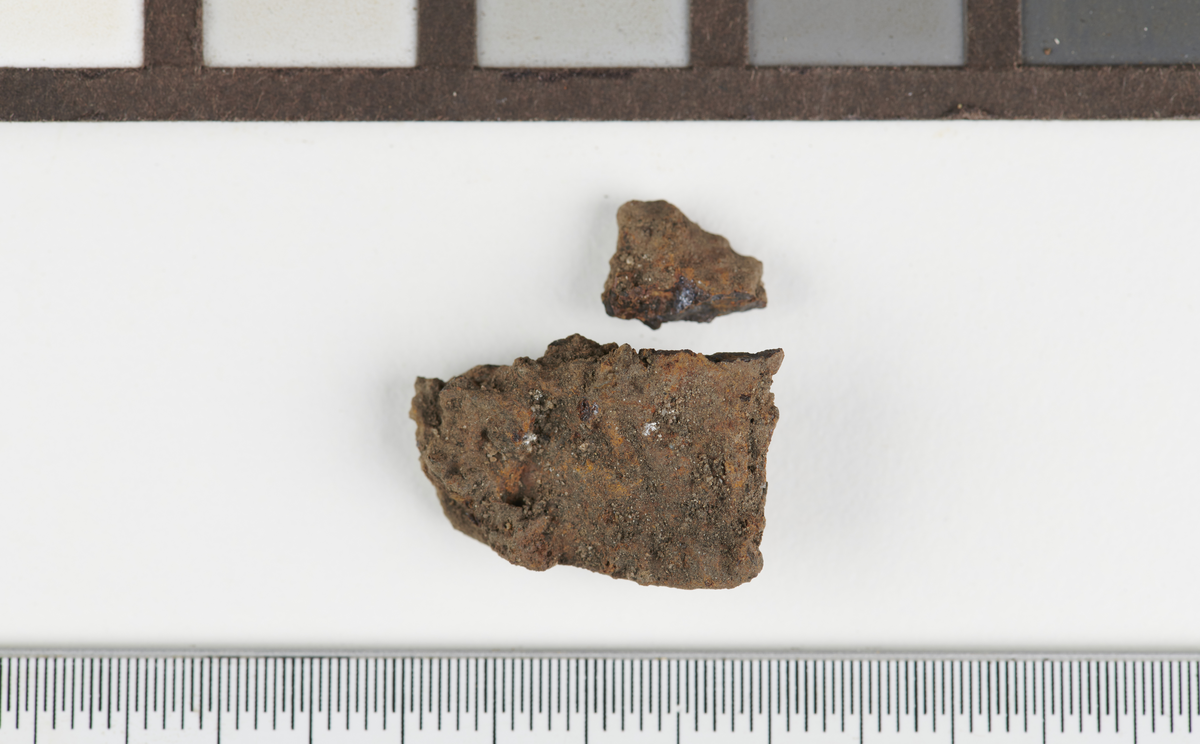 Flatt fragment av jern, i to deler. Mål: L: 2,5 cm. B: 1,8 cm. T: 0,7 cm. Vekt: 2,8 gram.
