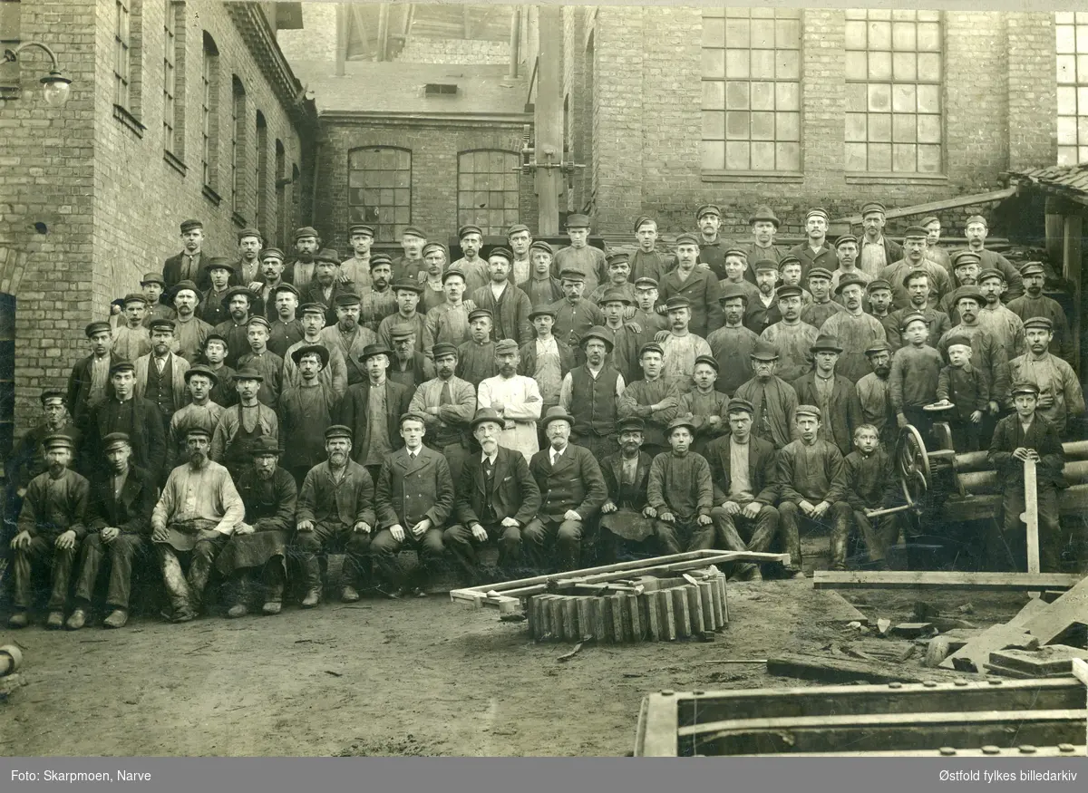 Antakelig et arbeidsmannskap fra Cathrineholm jernverk ved Tistedalsfossen i Halden, ca. 1890-1900. 

Cathrineholm produserte støpejernsprodukt, ovner og landbruksutstyr. Fra 1907 la de om drifta til emaljeverk som produserte skilt, sanitær- og kjøkkenutstyr.
