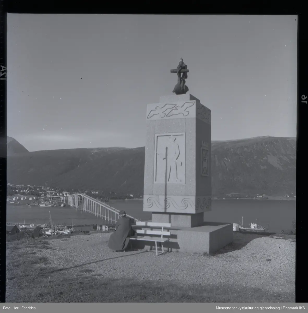 Monument ved "Tromsø Maritime skole". En kvinne sitter på en benk og ser ut over Tromsøysundet. I bakgrunnen ser man Tromsø-brua, fjell og bebyggelse.
