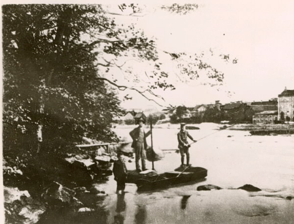 Laxmetare i Ätran, två män och en pojke. De har metspö, håv och en laxeka. På andra sidan ån syns delar av Falkenberg.