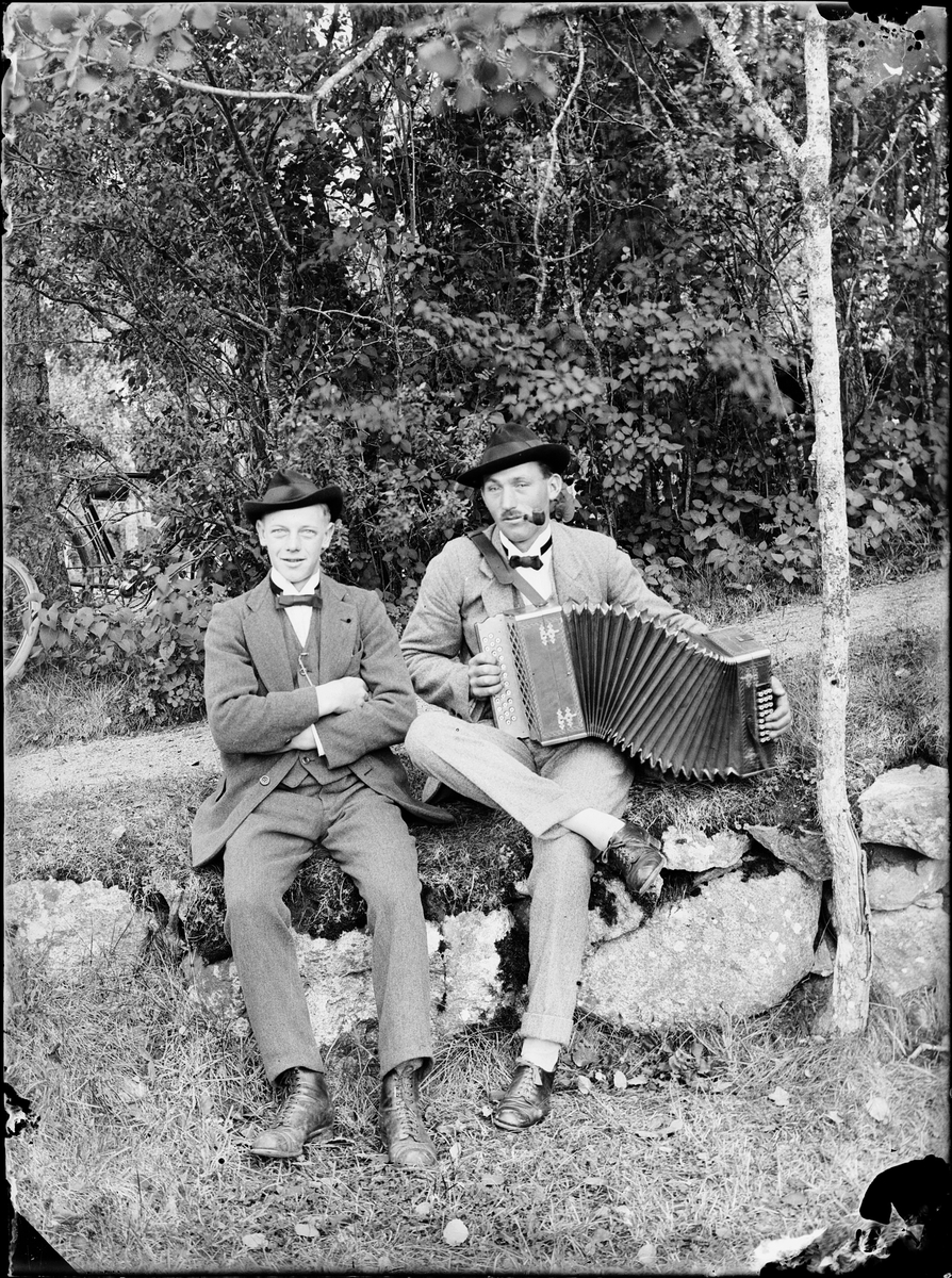 Två män sittandes på en stenmur, den ena spelar på ett dragspel, Alunda, Uppland