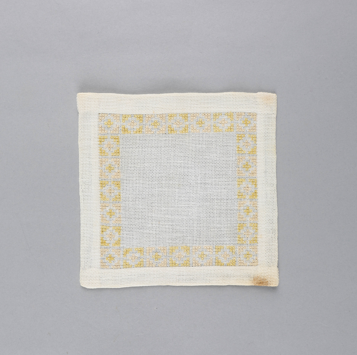 Tilnærmet kvadratisk dekkebrikke i hvitt tekstil med brodert bord langs brettekanten. Broderiet består av korssting av gul og rosa tråd i geometrisk mønster.
