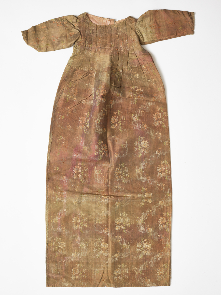 Dåpskjole i blomstret mønster. Fòret øverst med rosa bomull. Den er sannsynligvis sydd om av en eldre kjole.