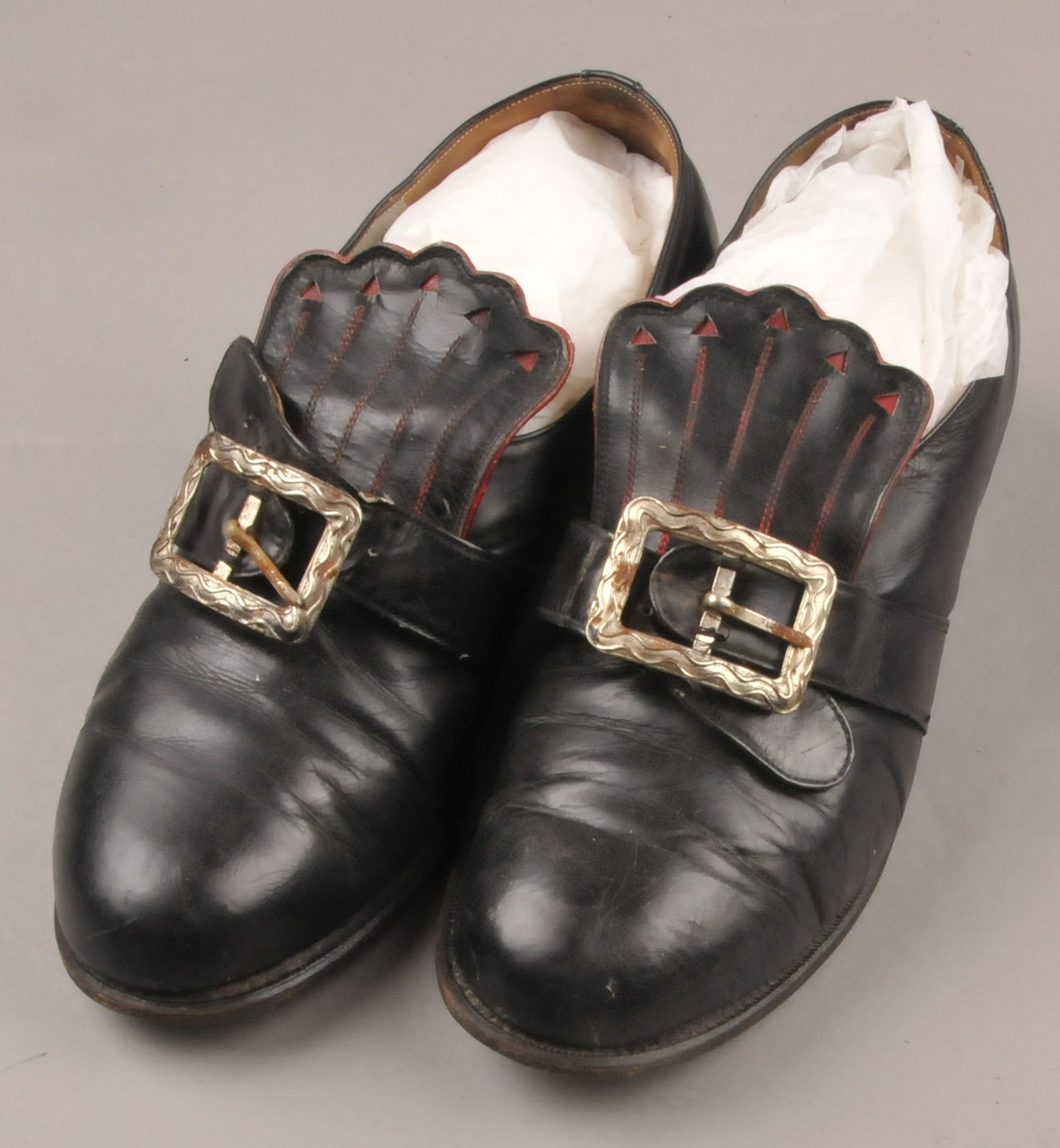 Svarte bunadsko med raud dekor på pløsa. Støpt metallspenne. Skoene har fått nye hælar, som er plugga med metallnaglar.