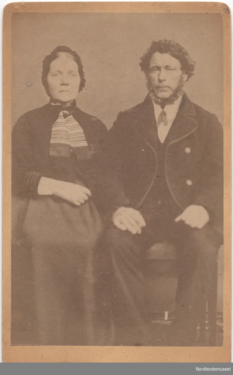 Portrett av et eldre par. Marie og Nils Grønbech. Paret sitter på stoler. Bildet er tatt utendørs foran et lerret.