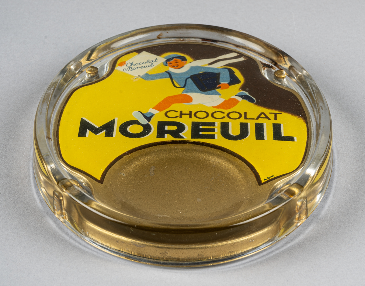 Myntbricka, oval, i glas, med guld, gul och svart bakgrund och springande pojke samt texten "CHOCOLAT MOREUIL" i svart.
