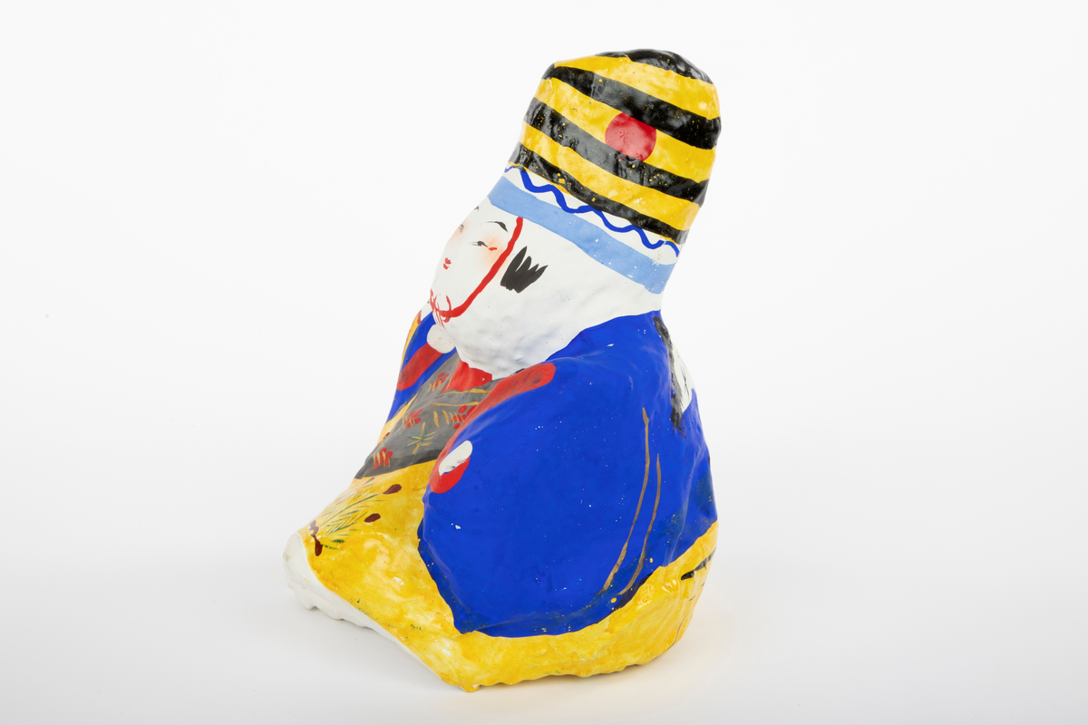 Mannsdukke i pappmasjé med stort hode og liten, kompakt kropp. Figuren har gul bukse, grå vest, blå jakke og en gul- og svartstripete hatt. På dukken er det malt flere lykkesymboler, blant annet en trane på ryggen.