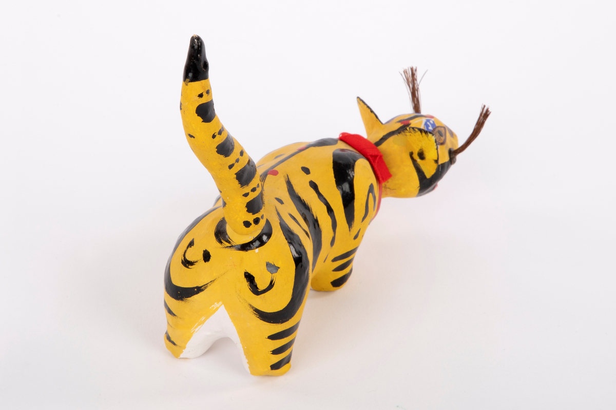 Tiger i pappmasjé med bevegelig hode. Tigeren er gul med svarte striper, har lang kropp med løs hale og et rødt bånd rundt nakken. Figuren har utstående ører, værhår og åpen munn med hvite tenner.