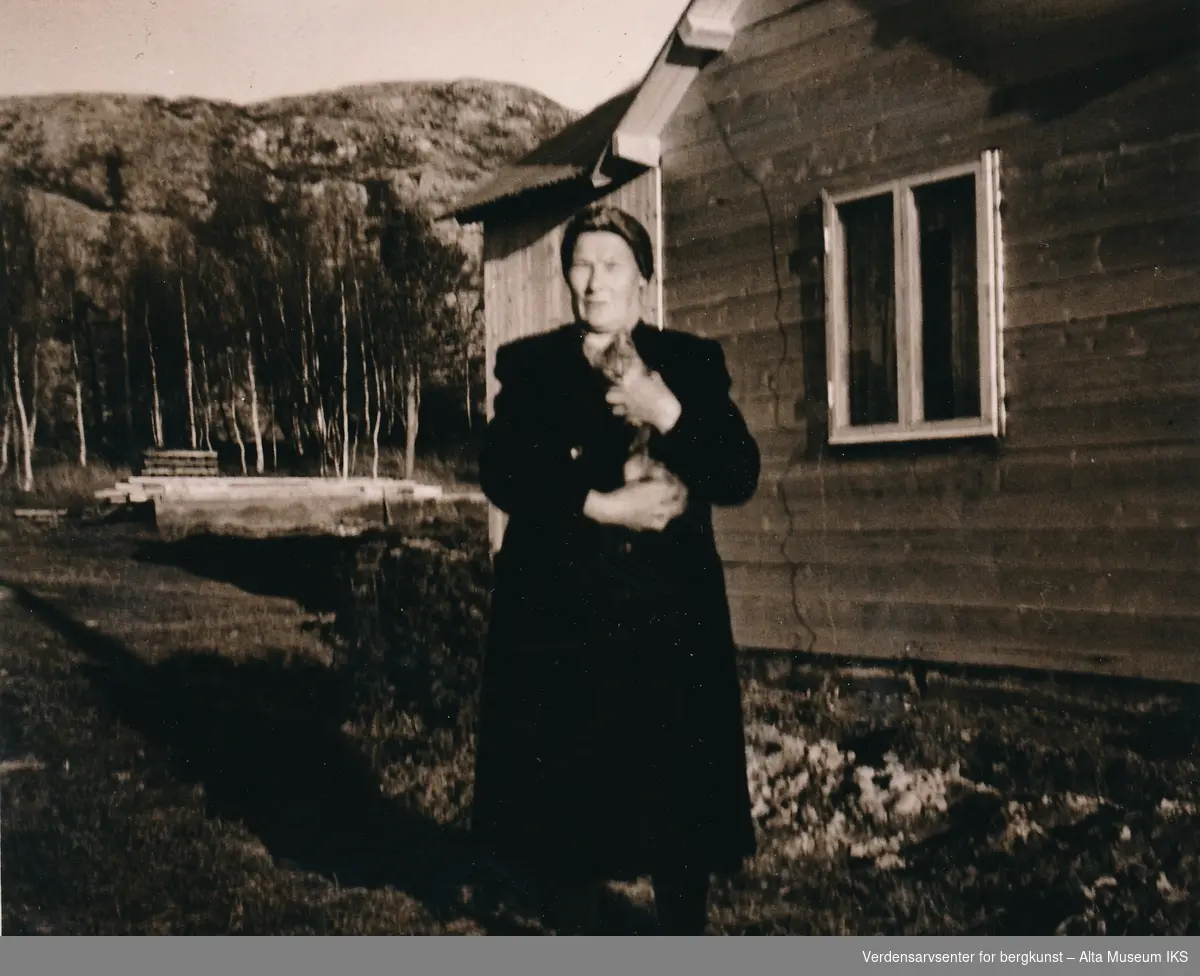 Kvinne med kattunge utenfor et hus med byggemateriale og fjell i bakgrunnen.