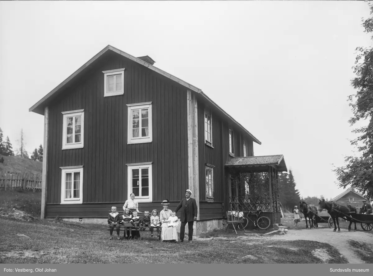 Vy mot ett bostadshus i två våningar i Berga, Mjösund. En familj med sex barn på en bänk framför och till höger syns ytterligare två personer med två hästar spända framför varsin vagn. Invid den lövade brokvisten står en cykel och ett ställ där de diskade mjölkkrukorna hänger på tork. Se även SuM-foto035104.