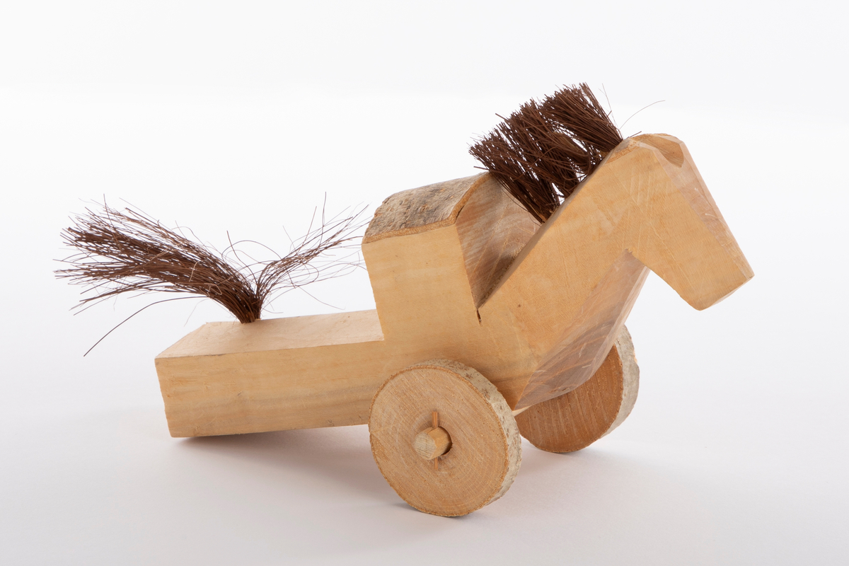 Hest på to hjul, skåret i et stykke ubehandlet tre. Det er skåret opp en forhøyning på ryggen; en sal. Rundt hjulene og på salen er det bark. Man og hale er laget av hamp.
